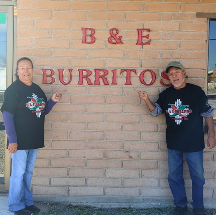 Founders of B & E Burritos standing outside the restaurant in Hatch, New Mexico, wearing logo shirts and smiling proudly by the brick wall sign.